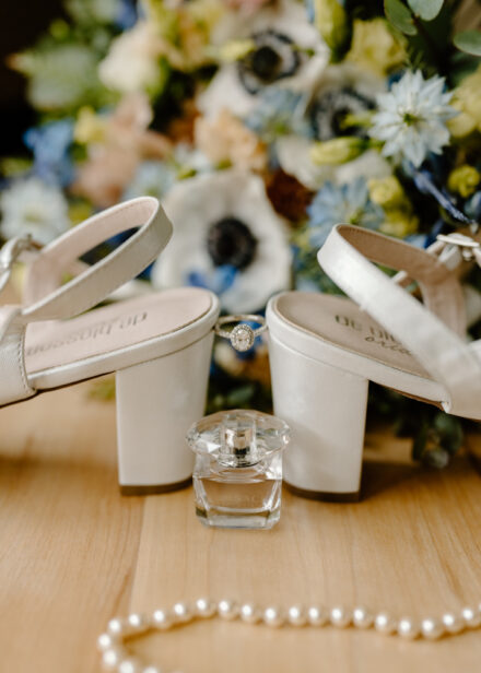 The bride's engagement ring is suspended between the heels of her shoes, flowers in the background.