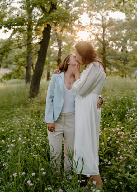 A queer couple kisses in a patch of wildflowers with the sun setting behind them.