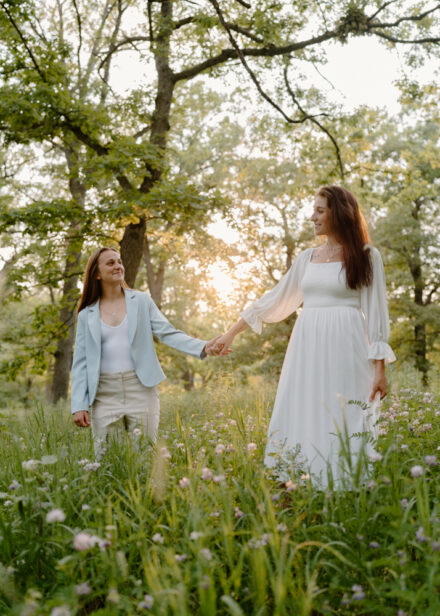 A queer couple holds hands in a patch of wildflowers with the sun setting behind them.