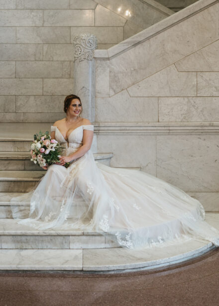 Connie smiles off camera as she sits on the white marble staircase in her wedding dress.