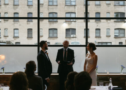 Beth and Matt stand with their officiant in front of the wall of windows in the private dining space.