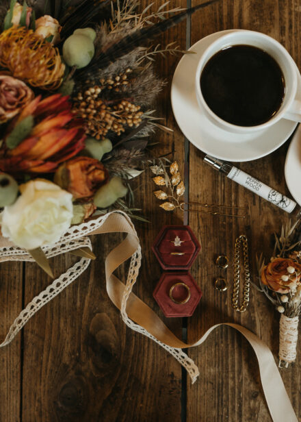 A detail shot of the orange and white bouquet, rings in a maroon velvet box, jewelry, and a cup of black coffee.