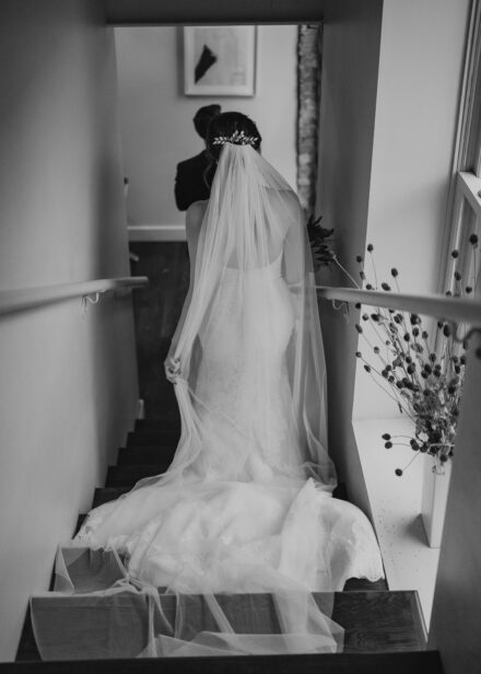 Beth walks down the stairs, veil and train swept dramatically behind her.
