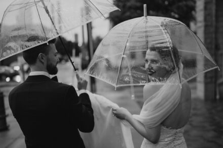 Beth peeks over her shoulder at the camera through her clear umbrella as she walks down the street with Matt in the rain.