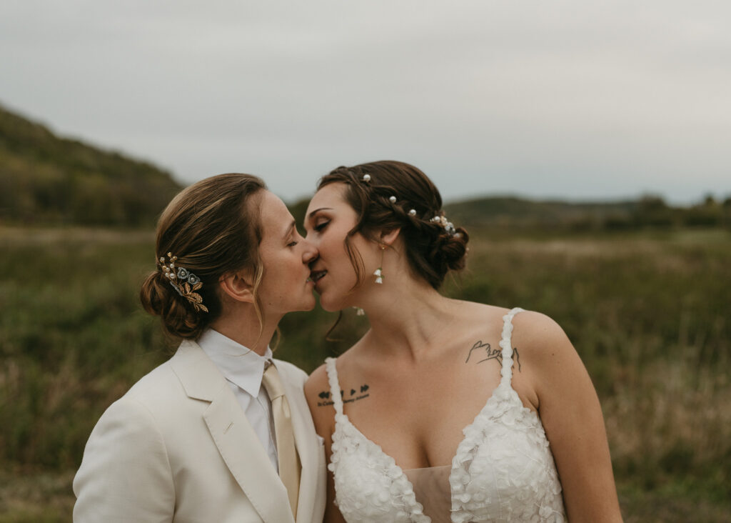 A lesbian couple kisses in front of a gold and green field on their wedding day