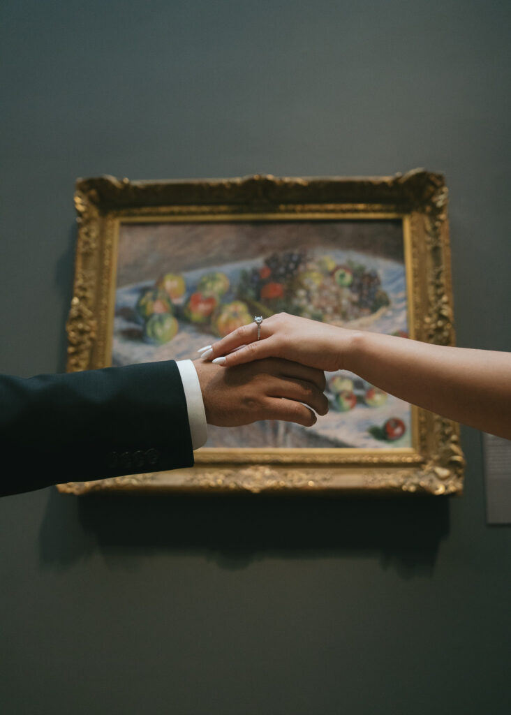 A close up of the couple's hands reaching toward each other in front of a still life painting.