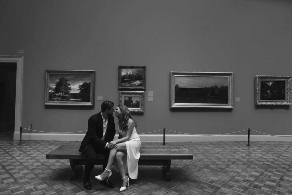 A couple leans in for a kiss on a bench in an art gallery.