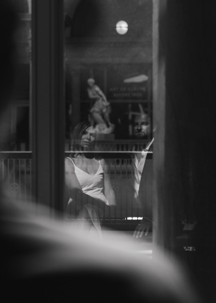 A couple's reflection is visible in the window as they sit on a museum bench.