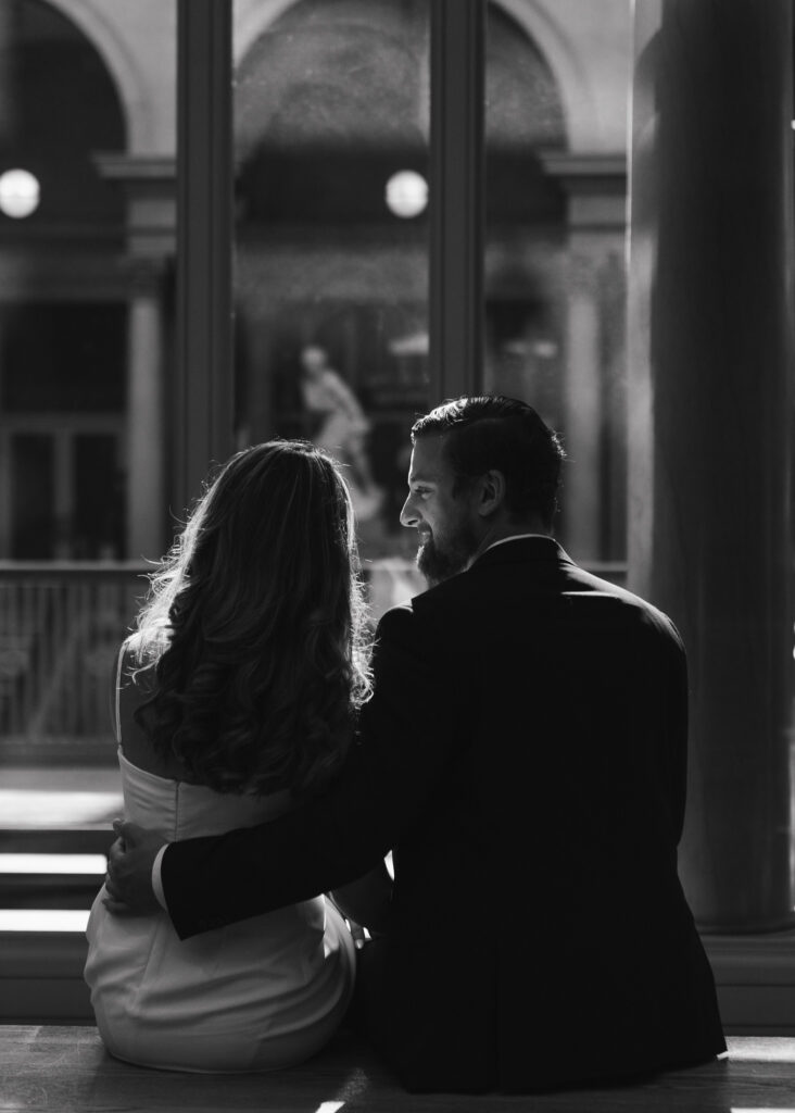 A man looks at his fiance while sitting on a museum bench, the sun glowing around them.