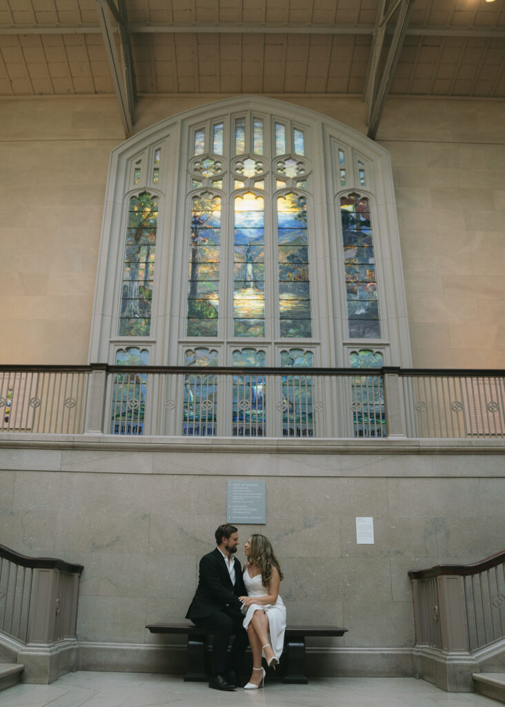 A couple sits on a bench in front of a stained glass window.