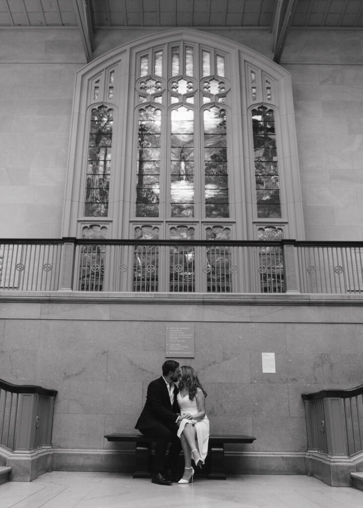 A couple kisses on a bench in front of a large stained glass window.