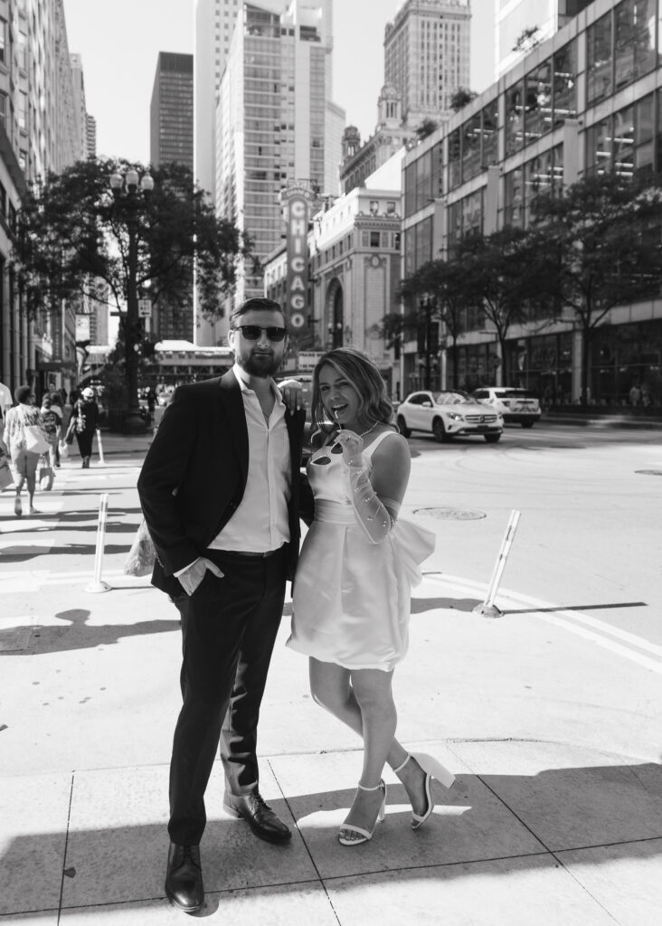 A couple looks seriously at the camera, the woman's sunglasses between her teeth.