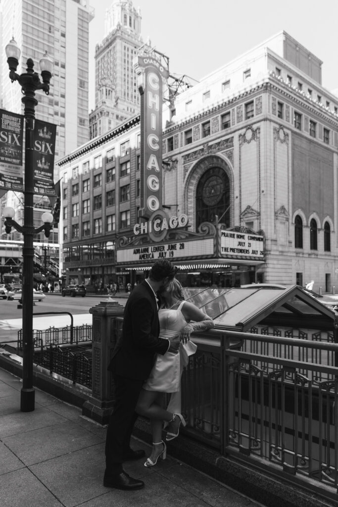 The couple kisses as they lean against an entrance to the L in front of the Chicago Theater.