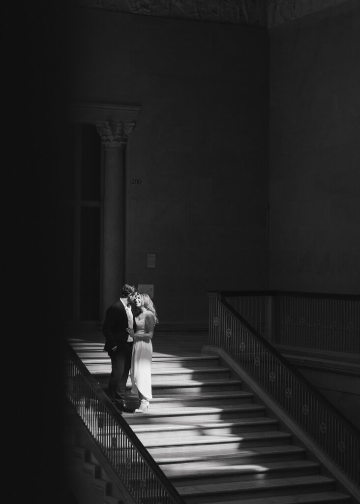 A couple leans in for a kiss in a patch of sunlight on the Grand Staircase in black and white.