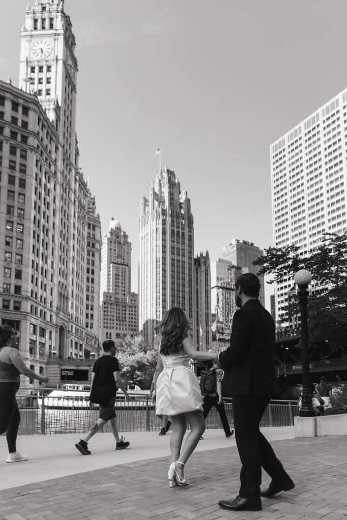 The couple holds hands, the woman walking away from the camera toward the river as people walk by.