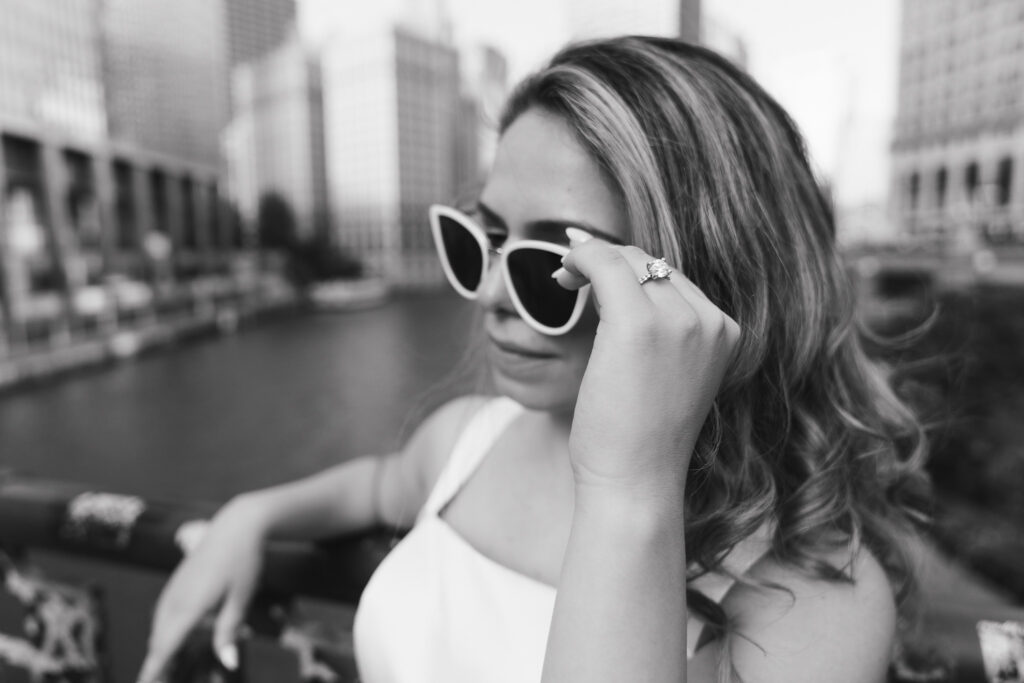 The woman holds the side of her sunglasses, engagement ring in focus.