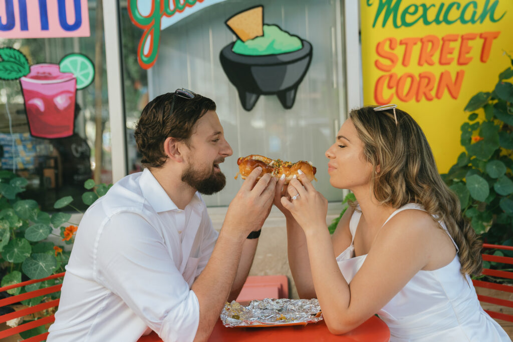 The couple eats a Chicago dog together on the colorful patio of a restaurant.