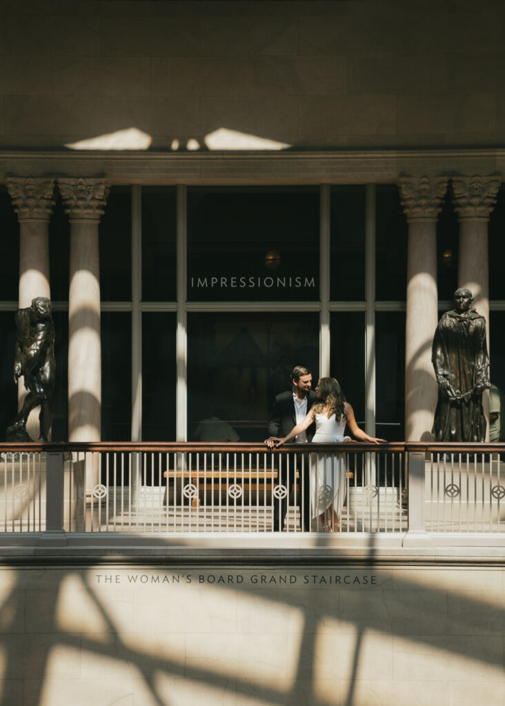 A couple leans on a balcony railing in front of a museum gallery