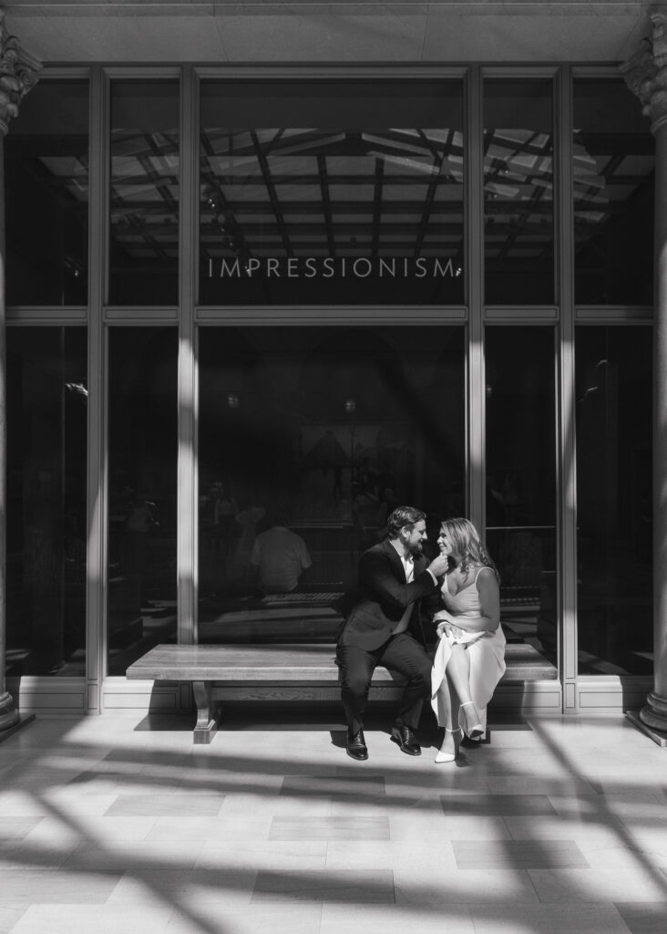 A man brings his hand up to his fiance's chin as they sit in a patch of sunlight on a bench.