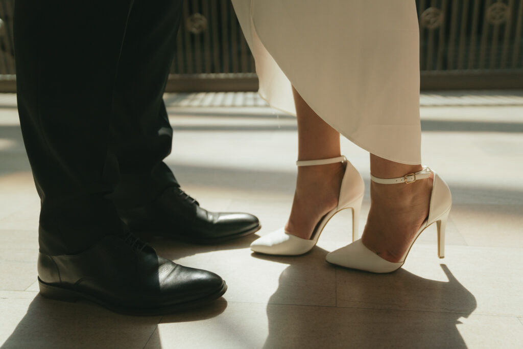 A close up of the couple's shoes, one in white strappy heels and one in black loafers.