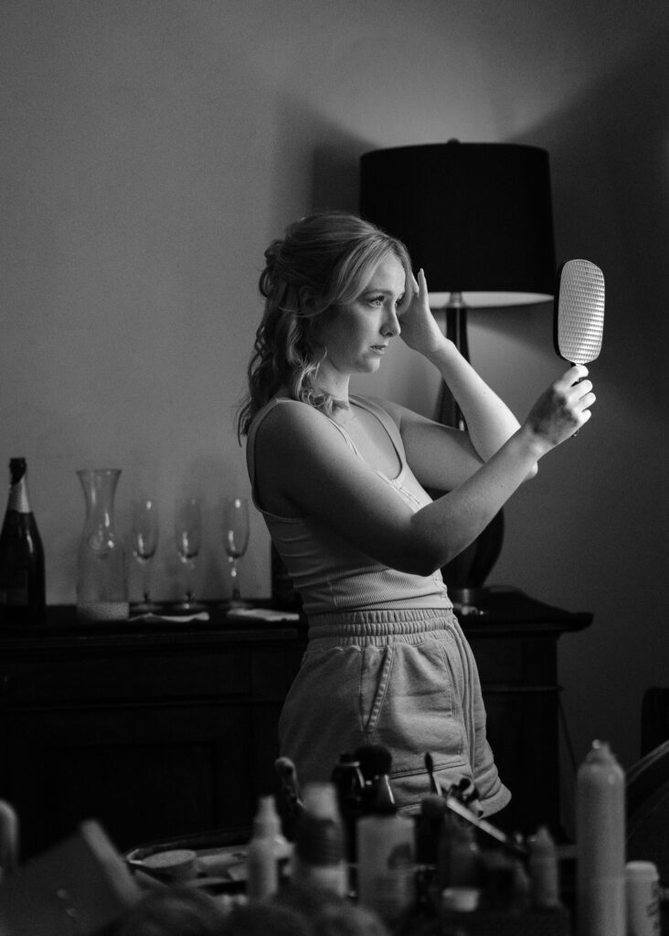 The bride fixes her hair in a handheld mirror in the middle of a messy room.