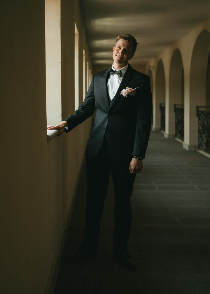 The groom smiles at the camera with his hand on a windowsill.