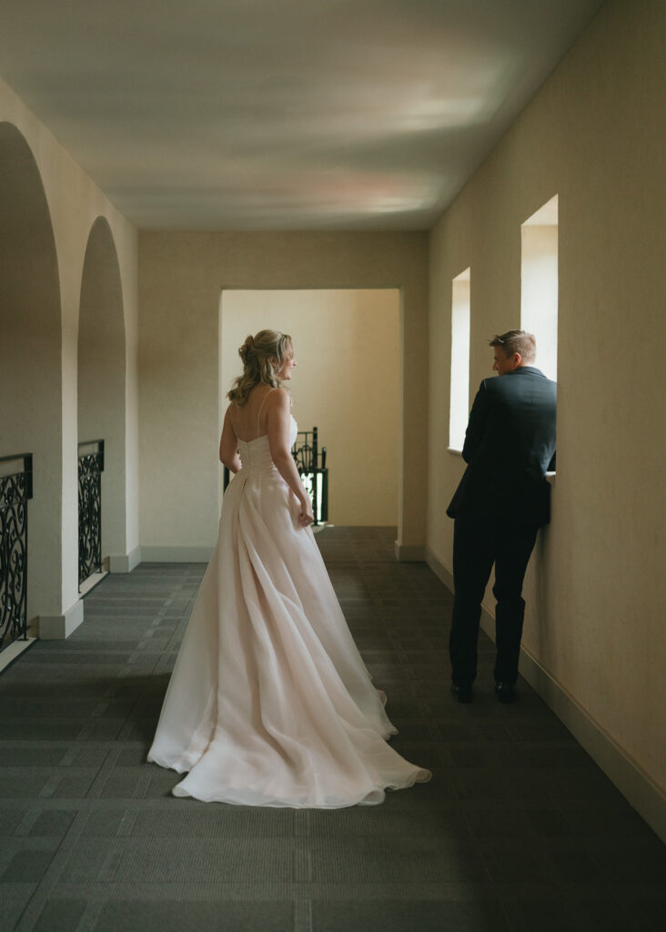 The bride walks down a long hallway with natural light toward the groom for their first look.