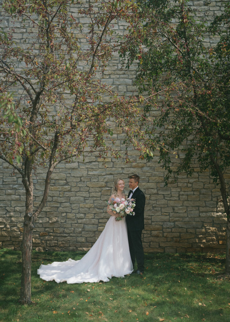 The bride and groom stand side by side in front of a stone wall, underneath a green and red tree.