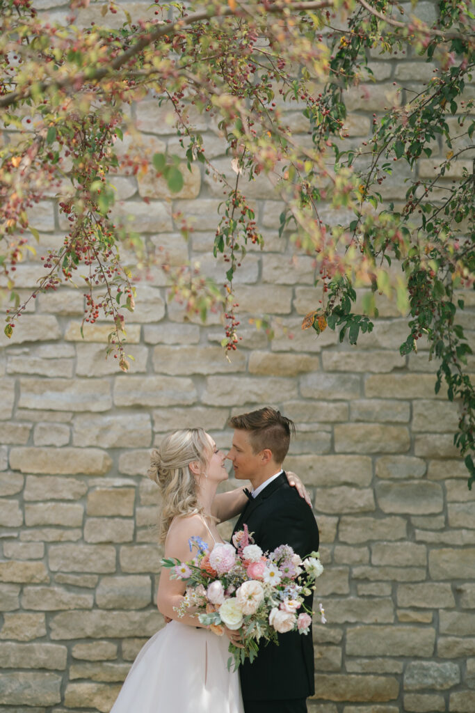 The couple leans in for a kiss in front of a stone wall, the bride holding her bouquet at her hip.