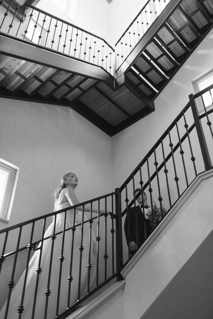 The bride and groom walk up a spiral staircase, the bride looking up at the stairs snd the groom looking back at her.