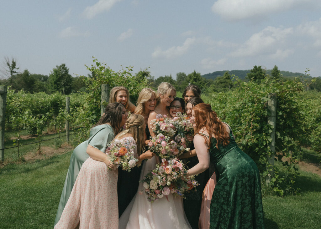 The bride is embraced by all of her bridesmaids as they stand in front of the vineyard.