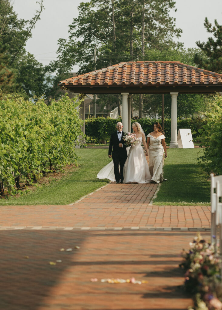 The bride walks down the aisle with both of her parents, between the rows of the vineyard.