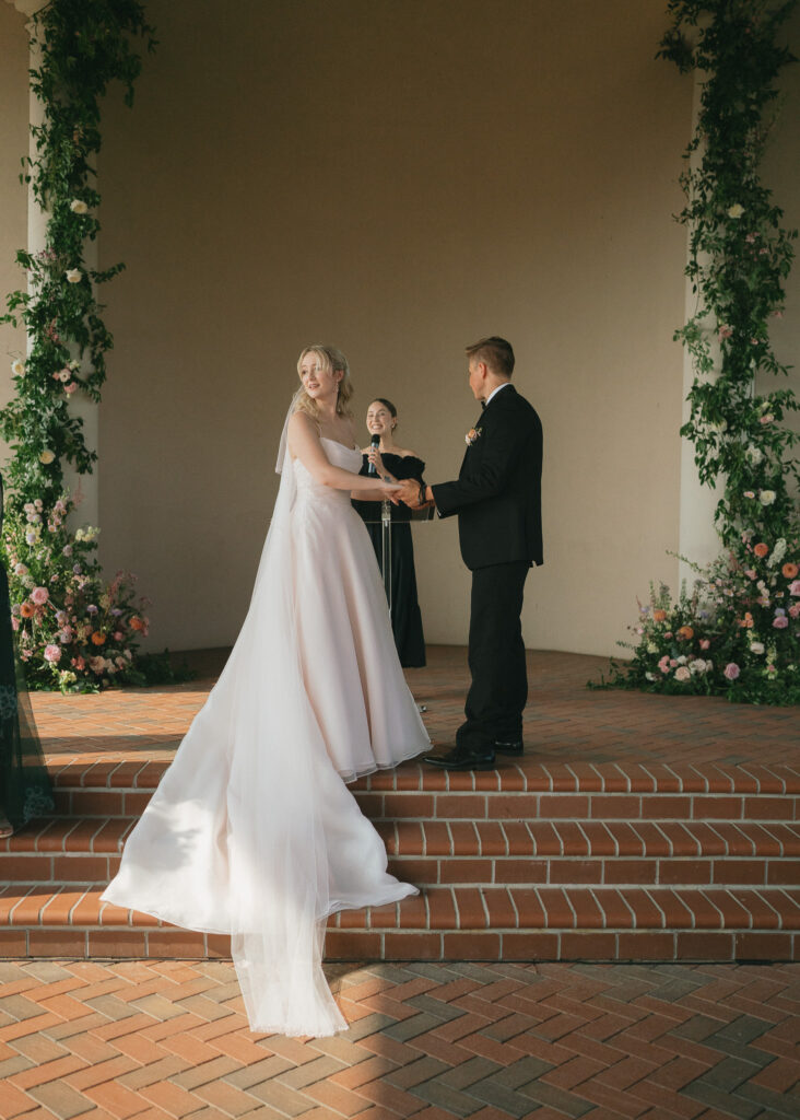 The bride and groom hold hands on the dais, between the floral arches.