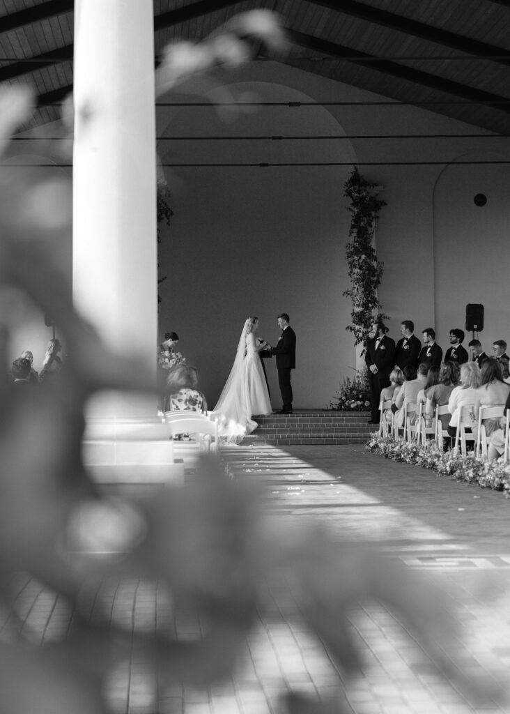 The couple holds hands during the wedding ceremony, vines from the vineyard in the foreground.