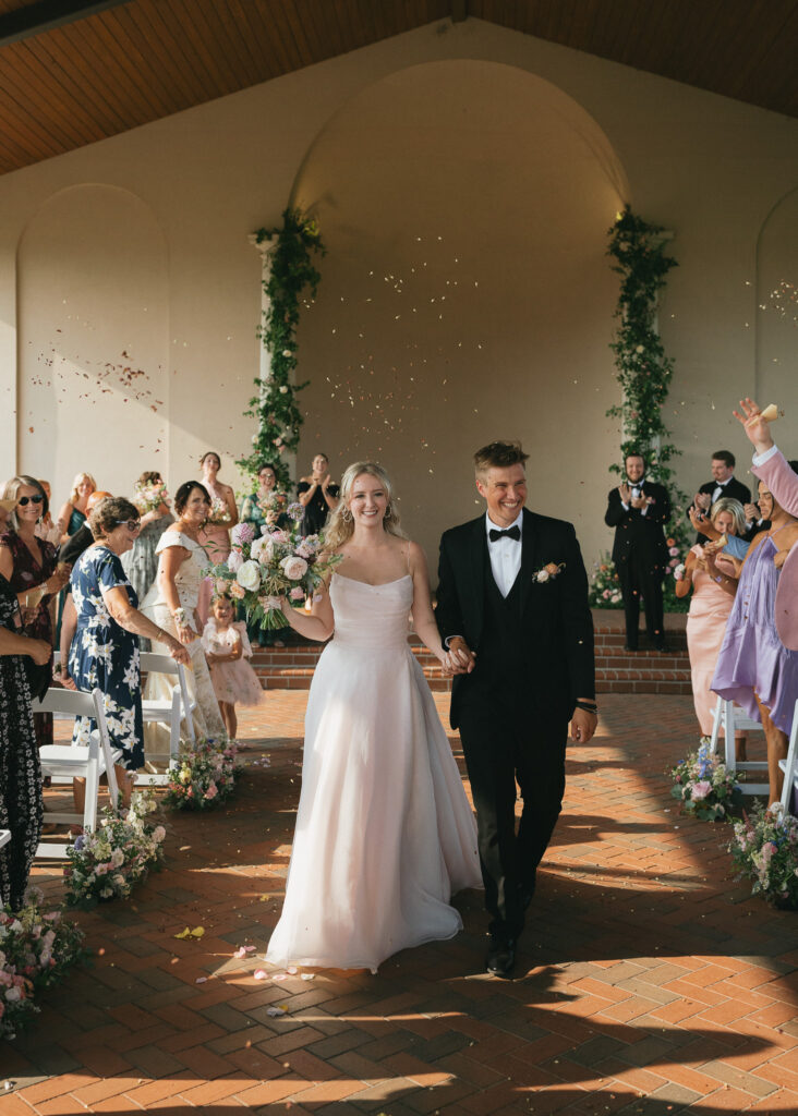 The couple smiles as they walk down the aisle, guests throwing dried flower petals over them.