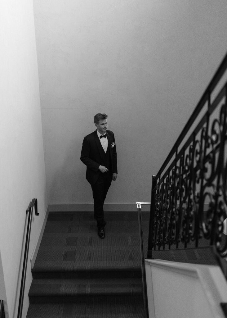 The groom waits at the bottom of the staircase for the bride before they enter the reception.