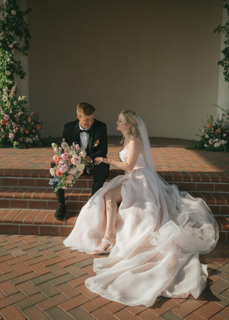 The couple sits on the brick steps of the vineyard pavilion, the groom holding the bouquet and the bride's dress cascading down the stairs.