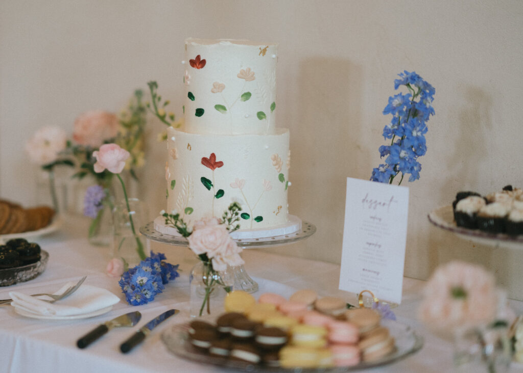 The dessert table filled with macarons, cupcakes, and a white, two-tiered cake with pressed flowers.