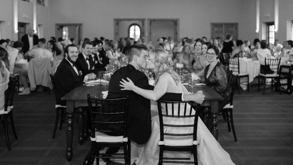 The couple kisses as they sit at the end of a long table, their wedding party smiling down the table at them.