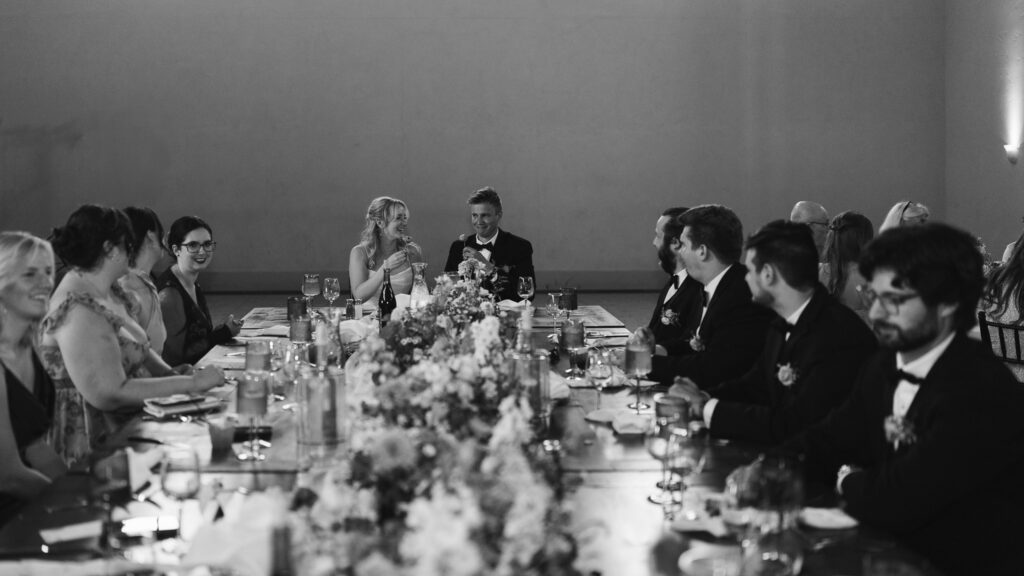 The couple and wedding party eat and drink along the sides of a long banquet table.