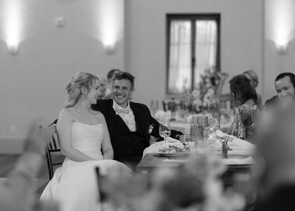 The bride smiles at the groom as he laughs during a speech.