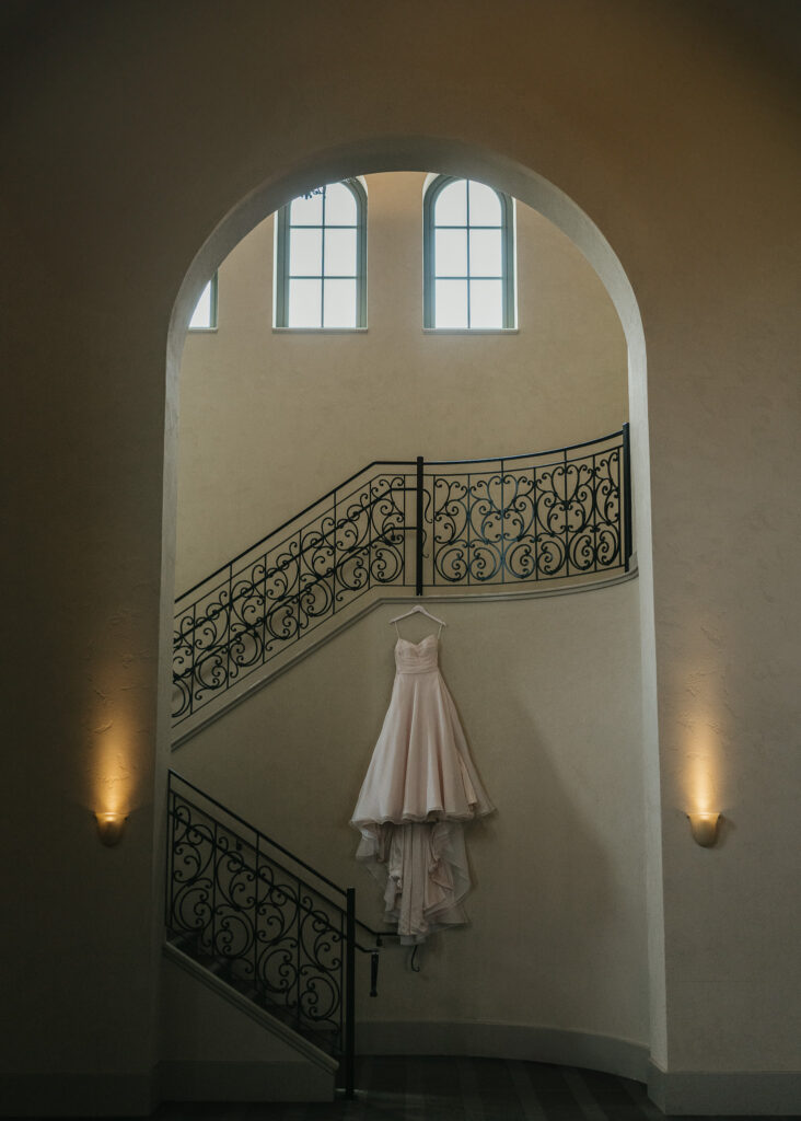 A wedding dress hangs from a wrought iron staircase, underneath a sweeping archway.