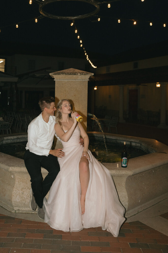 The bride drinks directly from a bottle of white wine, the groom's arms wrapped around her as they sit on the edge of the courtyard fountain.