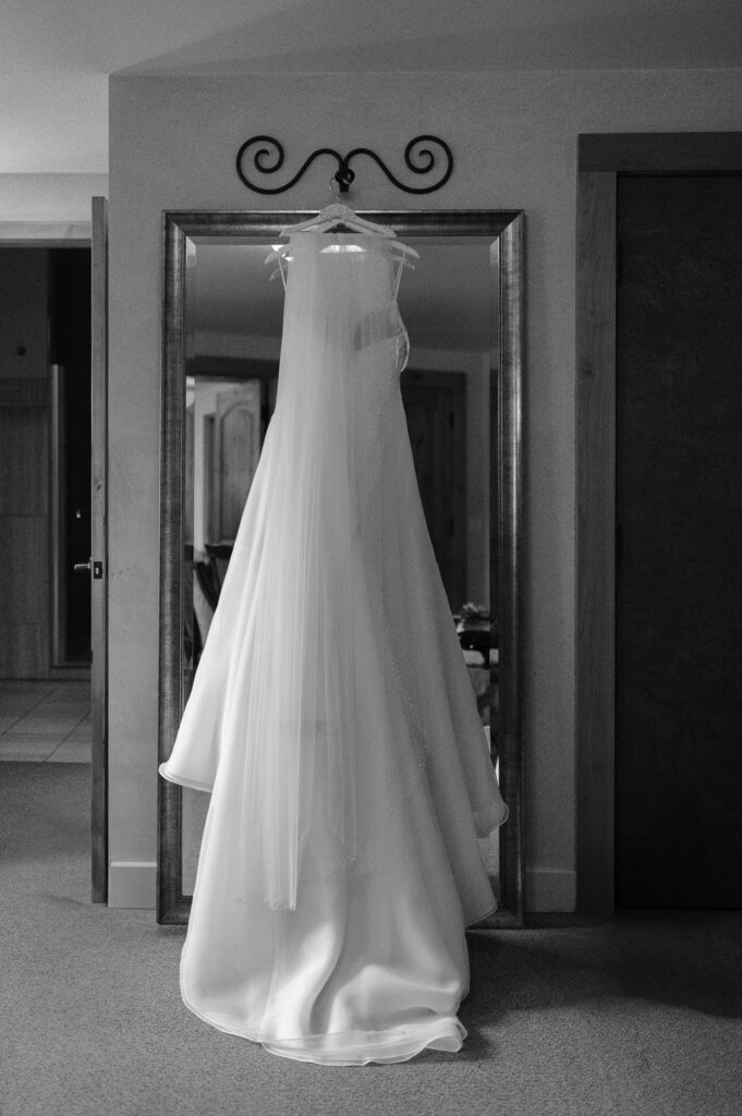 A black and white image of a wedding dress and veil hanging from a floor length mirror.