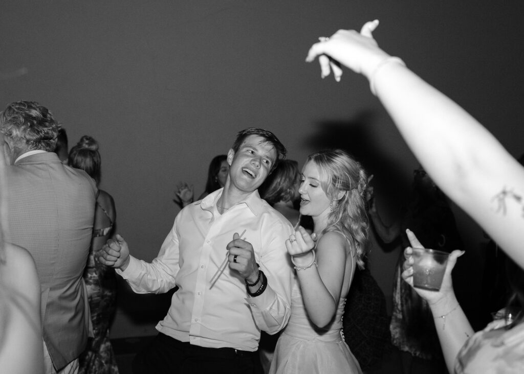 The couple dances together on the dance floor, a guests arm framing them in the foreground.